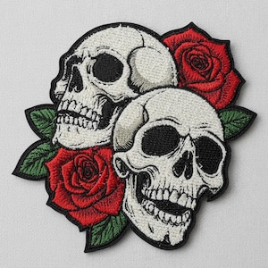 Może przedstawiać: Haftowana naszywka przedstawiająca dwie białe czaszki z czarnymi oczodołami i zębami, otoczone czerwonymi różami i zielonymi liśćmi. Naszywka ma czarną obwódkę, idealna do odzieży lub akcesoriów.