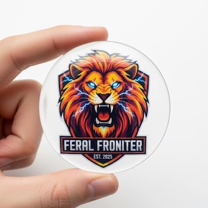 May include: A clear, round button featuring a fierce lion's head with a fiery orange and yellow mane, blue eyes, and lightning bolts. The text "FERAL FRONITER EST. 2025" is displayed below the lion graphic.