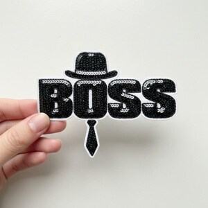 May include: A black sequined patch with the word "BOSS" in large, bubble letters. A black hat sits atop the "O", and a black tie hangs below. The patch is held by a hand against a white background.