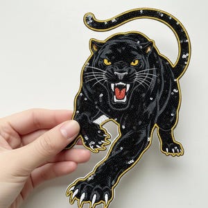 Op de afbeelding: Een zwarte panterpatch met gele ogen en een gouden rand. De patch is bedekt met pailletten en heeft een gedetailleerd ontwerp. De panter staat in een dynamische pose, met één poot uitgestrekt.