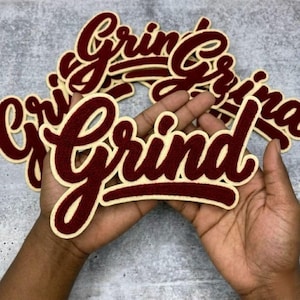 Puede incluir: Parches bordados de color granate y crema con la palabra "Grind" en fuente cursiva. Se ven varios parches, uno de ellos sostenido en el centro de la imagen. Los parches tienen una textura similar al fieltro y un estilo universitario clásico.