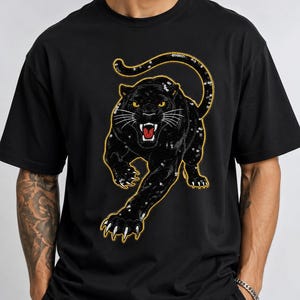 Puede incluir: Una camiseta negra de manga corta con un gran gráfico central de una pantera negra rugiendo. La pantera está detallada con lentejuelas negras, ojos amarillos, una boca roja y un contorno de color dorado, en una postura de salto.