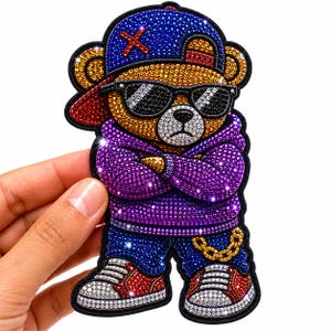 Peut inclure: Un écusson décoratif représentant un ours portant une casquette bleue avec un X rouge, des lunettes de soleil noires, un sweat à capuche violet, un pantalon bleu avec une chaîne dorée et des baskets rouges et blanches, orné de strass scintillants.