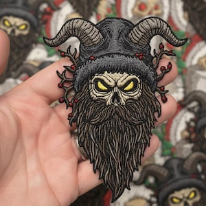 Könnte beinhalten: Ein gestickter Aufnäher mit einem Totenkopf mit langem braunen Bart, der eine schwarze Mütze mit zwei großen geschwungenen Hörnern trägt. Das Design enthält kleine braune Zweige mit roten Beeren an beiden Seiten und leuchtend gelbe Augen.
