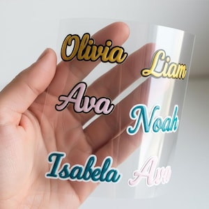 May include: A clear sheet with several names in a cursive font. The names include "Olivia" in gold, "Liam" in gold, "Ava" in pink, "Noah" in teal, "Isabela" in teal, and "Ava" in pink. The names have a 3D effect.