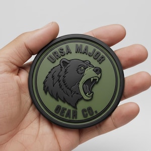 May include: A round patch with a black border and a green center featuring a roaring bear graphic. The text "URSA MAJOR" arches above the bear, and "GEAR CO." is below. The bear is black with a green mouth.