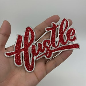 Op de afbeelding: Een rode hanger met het woord "Hustle" in een cursief lettertype. De letters zijn gevuld met rode strass-steentjes en omlijnd met heldere strass-steentjes. De hanger wordt in een hand gehouden.