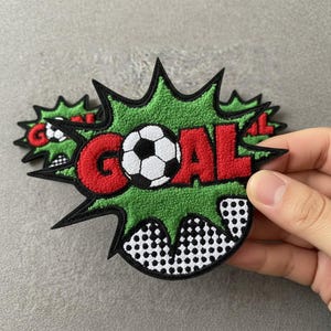 May include: A green, red, black, and white patch with the word "GOAL" and a soccer ball. The patch is in a comic book style, with a green textured background and a black outline.