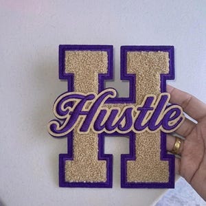 Puede incluir: Un parche morado y dorado estilo universitario con la forma de la letra "H". La letra está delineada en morado con un centro texturizado dorado. La palabra "Hustle" está escrita en cursiva en el centro de la letra.
