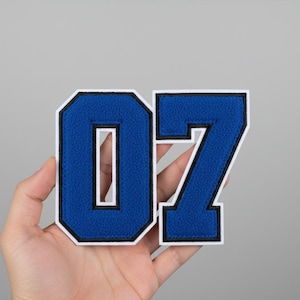 Puede incluir: Un parche azul y blanco con el número 07. El número es de estilo universitario con un borde blanco y un contorno negro. El número se sostiene en una mano.