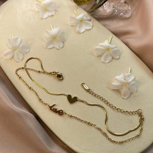 May include: A gold-colored necklace with a heart pendant and the word "love" is displayed on a cream-colored tray, alongside white flowers and gold bangles. The jewelry is set against a backdrop of draped, peach-colored fabric.