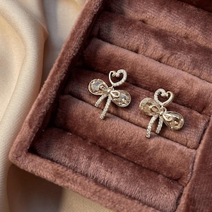 Può includere: Un paio di orecchini a lobo color oro con una parte superiore a forma di cuore e un centro simile a un cristallo. Gli orecchini sono esposti in un portagioie di velluto rosa scuro, su uno sfondo di tessuto di raso beige chiaro.