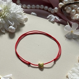 Peut inclure: Un bracelet en fil rouge avec un petit cœur doré. Le bracelet est présenté sur une surface crème, entouré de fleurs blanches, de perles et d'un fond en tissu rose. Un bijou délicat et élégant.
