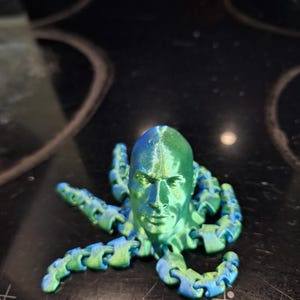 May include: A 3D-printed octopus figure with a human face, in a blue and green gradient. The articulated tentacles are segmented. The head has detailed facial features.