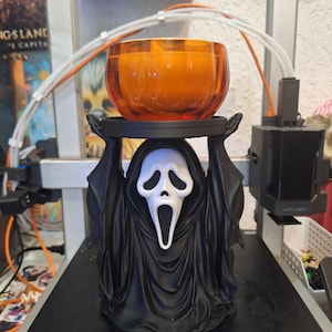 May include: A black 3D-printed figure of a cloaked character with a white mask featuring a long, open-mouthed expression. The figure holds a circular black tray above its head, which supports a round, translucent orange glass candle holder.