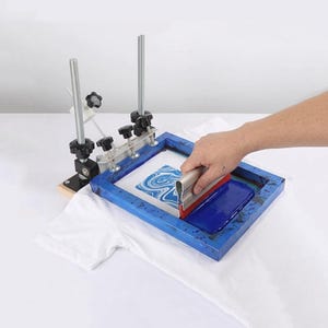 Puede incluir: Un marco de serigrafía azul con una rasqueta metálica aplicando tinta azul sobre una camiseta blanca. El aparato cuenta con varillas metálicas verticales, perillas de ajuste negras y un sistema de bisagras montado sobre una base de madera.