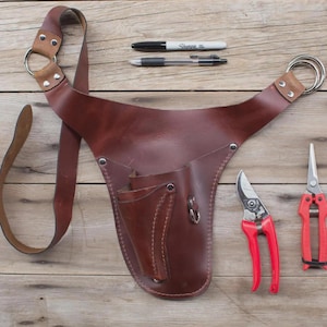 Op de afbeelding: Een bruine leren tuingereedschapsriem met een schouderriem en metalen ringen. Twee rood-handvat snoeischaar en twee pennen zijn ook zichtbaar. De riem heeft zakken en een lus voor gereedschap.