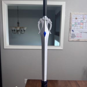 May include: A silver-colored sword with a black twisted handle and a silver pommel. The crossguard features curved silver accents and a central blue diamond-shaped inlay. The sword is mounted vertically on a dark purple circular base with floral details.