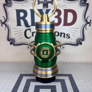 May include: A cylindrical green lantern prop featuring a metallic gold ring at the top, a central green circular emblem with a square cutout, and horizontal ribbed detailing. The object has gold and blue-grey bands at the top and bottom.