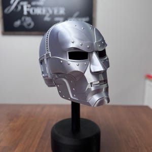 May include: A metallic silver full-face mask featuring a rigid, angular design with prominent rivet details along the cheekbones and forehead. The mask includes rectangular eye openings and a segmented jaw section, mounted on a black cylindrical stand.