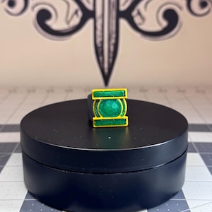 May include: A ring featuring a rectangular yellow frame with a circular green center emblem. The design includes a raised green circle within the yellow border, creating a distinct geometric look. The ring is displayed on a black circular stand.