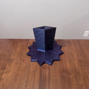 Puede incluir: Un soporte impreso en 3D de color morado oscuro con una columna central cuadrada y hueca que presenta intrincados patrones en relieve similares a circuitos, montado sobre una base ancha y plana en forma de estrella con surcos circulares.