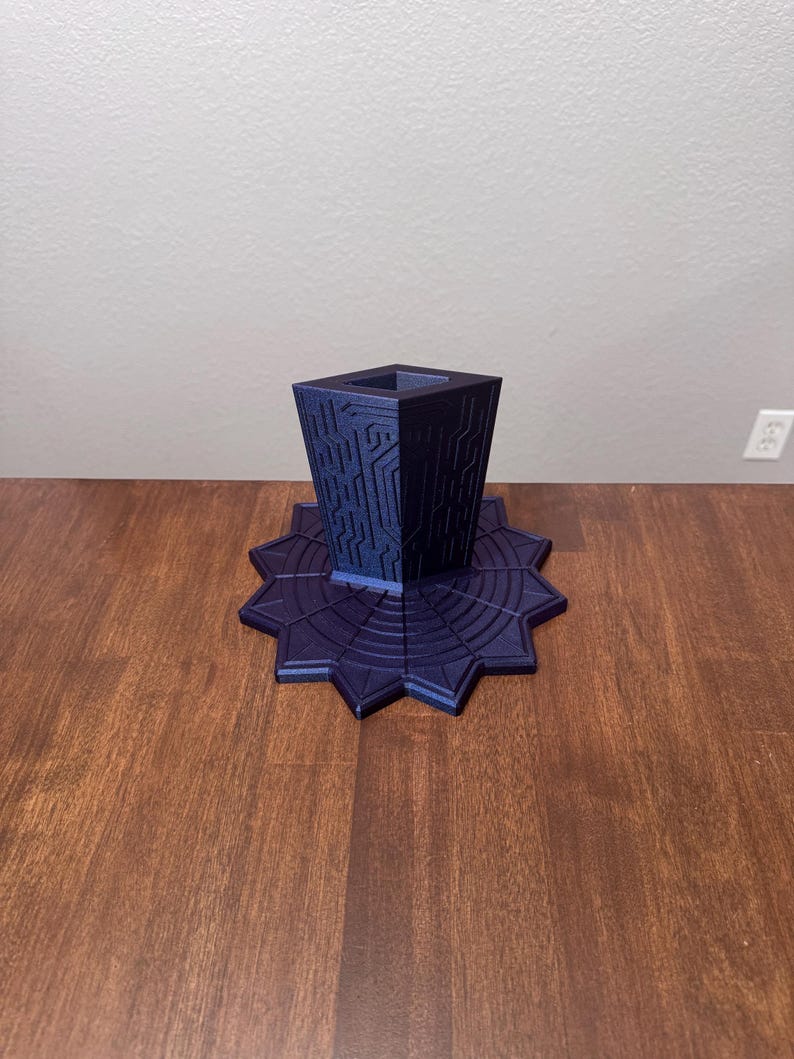 Puede incluir: Un soporte impreso en 3D de color morado oscuro con una columna central cuadrada y hueca que presenta intrincados patrones en relieve similares a circuitos, montado sobre una base ancha y plana en forma de estrella con surcos circulares.