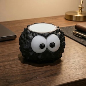 May include: A black, round decorative object with large, cartoonish white eyes and a white interior. The object sits on a wooden desk, alongside a laptop, notebook, and pen. The object's exterior has a textured, spiky appearance.