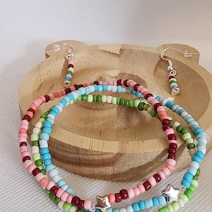 Könnte beinhalten: Bunte Perlenarmbänder und Ohrringe. Die Armbänder haben eine Mischung aus rosa, blauen, grünen und weißen Perlen mit silbernen und goldenen Sternanhängern. Die Ohrringe haben passende Perlen. Der Schmuck wird auf einem Holztablett präsentiert.