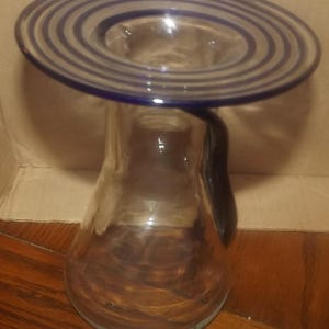 May include: A clear glass pitcher with a wide, flat rim featuring concentric dark blue circular bands. The pitcher has a tapered body and a dark blue handle attached to the side, extending from the base of the rim down to the mid-section.
