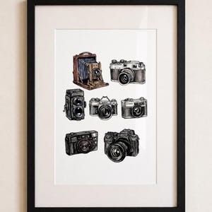 May include: A framed print featuring watercolor illustrations of various vintage cameras. The cameras are depicted in shades of brown, black, and silver, showcasing different designs and styles. The artwork is set against a white background.