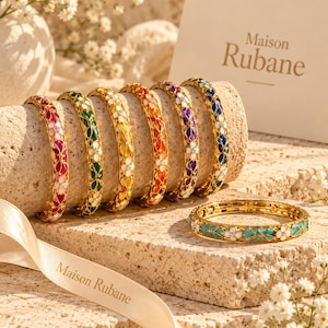 Pode incluir: Uma coleção de pulseiras de metal dourado com padrões florais de esmalte colorido em rosa, verde, amarelo, laranja, roxo, azul e turquesa. Cada pulseira apresenta um design floral repetitivo com centros brancos. Um cartão e uma fita exibem o texto Maison Rubane.