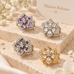 May include: Four flower-shaped rings featuring marquise-cut stones in purple, clear, grey, and clear with a gold setting. Each ring has a central round stone surrounded by petals and small accent stones. A card reads Maison Rubane.