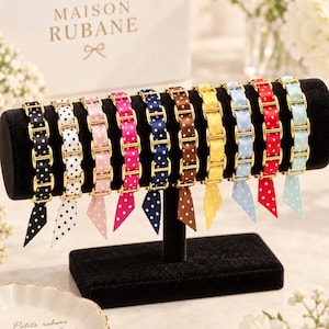 Pode incluir: Uma coleção de dez pulseiras exibidas num suporte de veludo preto. Cada pulseira apresenta uma fita de bolinhas em várias cores—preto, branco, rosa, magenta, azul-marinho, castanho, amarelo, azul-claro, vermelho e menta—entrelaçada com elos metálicos dourados.