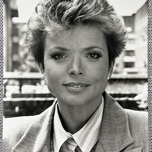 Pode incluir: Retrato a preto e branco de uma mulher com cabelo curto e penteado. Ela usa um blazer de cor clara sobre uma camisa de colarinho listrada. A imagem é um close-up, focando em seu rosto e parte superior do corpo.