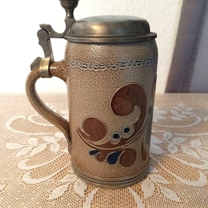 May include: A vintage ceramic beer stein with a hinged metal lid and handle. The stein is tan with brown and blue decorative swirls. The lid and handle are a dark metal color. The stein is sitting on a white lace tablecloth.
