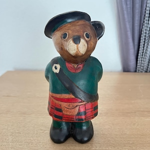 Op de afbeelding: Een handgesneden houten beeldje van een beer met een donkergroene baret, een bijpassend groen jasje en een rood-zwarte geruite kilt. De beer heeft een bruin gezicht, zwarte ogen en een zwarte neus, met een zwarte riem over de borst.