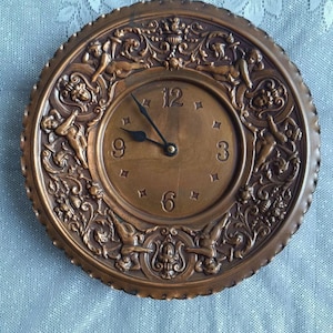 May include: A circular, bronze-toned wall clock with detailed relief decoration. The clock face displays Roman numerals and square markers. The outer edge is adorned with cherubic figures and floral designs. The hands are black.