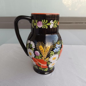 Könnte beinhalten: Ein schwarzer Keramikkrug mit handgemaltem Blumenmuster aus weißen Gänseblümchen, roten Mohnblumen, blauen Vergissmeinnicht und gelben Weizenähren. Der Krug hat eine glänzende Oberfläche, einen geschwungenen Henkel und einen leuchtend orangen Rand.