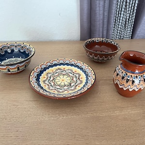 Pode incluir: Um conjunto de quatro peças de cerâmica com um padrão marmoreado em azul escuro, castanho e creme. A coleção inclui uma taça funda, um prato raso com um motivo floral central, uma taça castanha mais pequena e um jarro com pega.