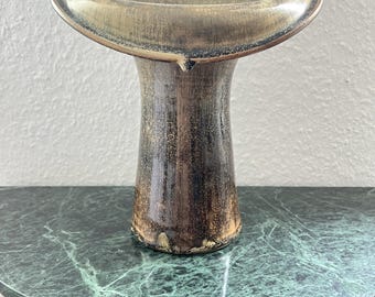 May include: A ceramic vase with a wide, flared, oval-shaped rim and a cylindrical body. The surface features a mottled, textured glaze in shades of dark brown, black, and muted gold, creating a weathered, metallic appearance throughout the piece.