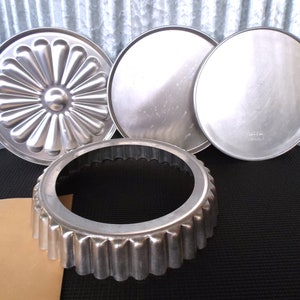 May include: A collection of vintage aluminum baking pans. Includes a fluted tart pan, two round pans, and a decorative pie pan with a sunburst design. The pans are arranged on a black surface with a corrugated metal background.