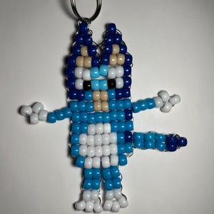 Puede incluir: Un llavero hecho a mano con un personaje de perro azul hecho de cuentas de colores. El perro tiene una combinación de colores azul, blanco y beige, con un llavero plateado en la parte superior. Los brazos y las piernas están extendidos, dando al personaje una apariencia juguetona.