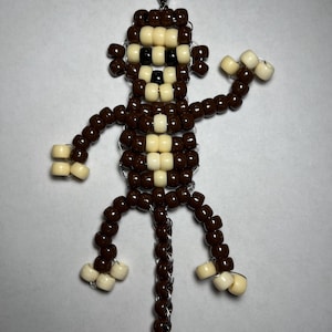 May include: A beaded monkey charm with a silver-colored clasp. The monkey is made of brown and cream-colored beads, with a black face. The arms and legs are outstretched, and the charm is likely for a keychain or accessory.