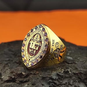 Op de afbeelding: Een gouden ring met een centraal schildembleem met een mijter en religieuze symbolen, omringd door kleine paarse stenen. De tekst luidt SEAL OF THE PRIMATE & PATRIARCH METRO. ABP. ERSKINE WILLIAMS, SR. en aan de zijkant SEAL OF THE met een sleutel.