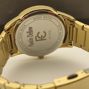 Op de afbeelding: Een goudkleurig polshorloge met een metalen schakelband. De achterkant van de horlogekast is zilverkleurig en bevat de tekst Austin DeBow, FC, 011772C, Nano Metal, 3ATM Water Resistant en Stainless Steel Case Back gegraveerd op het oppervlak.