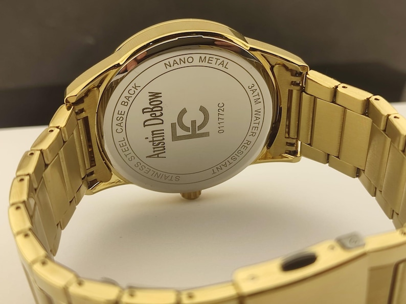 Op de afbeelding: Een goudkleurig polshorloge met een metalen schakelband. De achterkant van de horlogekast is zilverkleurig en bevat de tekst Austin DeBow, FC, 011772C, Nano Metal, 3ATM Water Resistant en Stainless Steel Case Back gegraveerd op het oppervlak.