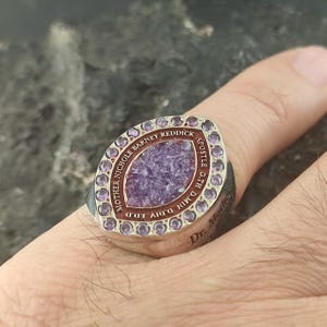 May include: A silver-toned ring featuring a marquise-shaped center with a textured purple stone. The center is surrounded by a border of small, round purple stones and an outer rim engraved with the text MOTHER NICHOLE BARNEY REDDICK APOSTLE D.TH D.MIN D.DIV ED.D.