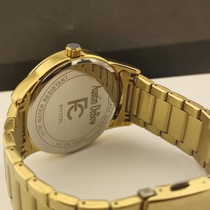 Op de afbeelding: Een goudkleurig metalen polshorloge met een schakelarmband. De achterkant van de horlogekast is zilverkleurig en bevat de tekst Austin DeBow, de letters FC, het nummer 011772C en de woorden 3ATM WATER RESISTANT en NANO METAL.