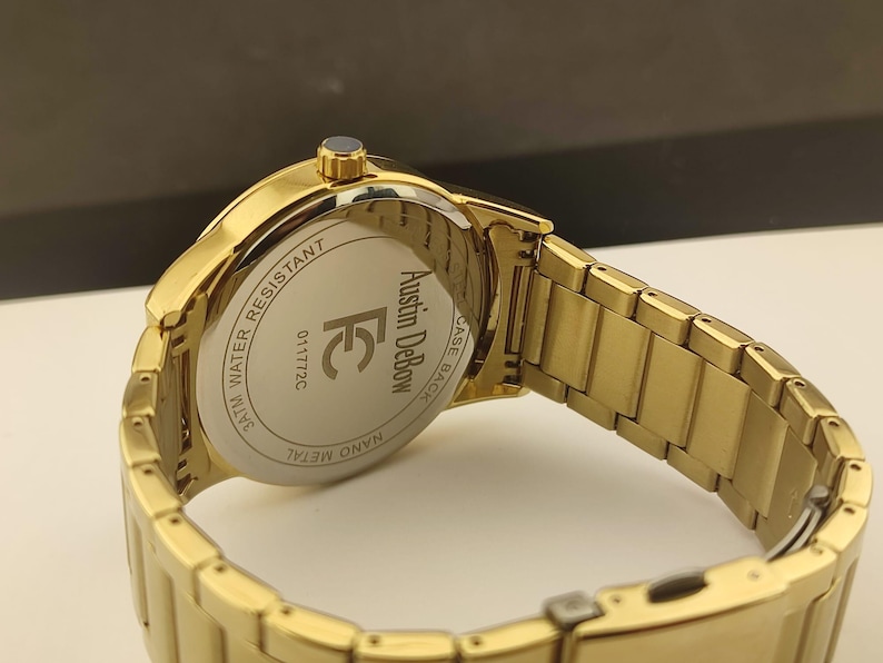 Op de afbeelding: Een goudkleurig metalen polshorloge met een schakelarmband. De achterkant van de horlogekast is zilverkleurig en bevat de tekst Austin DeBow, de letters FC, het nummer 011772C en de woorden 3ATM WATER RESISTANT en NANO METAL.